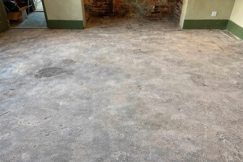 Ogołocone wnętrze po pracach rozbiórkowych: odsłonięta cegła kominka, surowa podłoga z widocznymi śladami po usuniętych elementach, zielone listwy przypodłogowe.