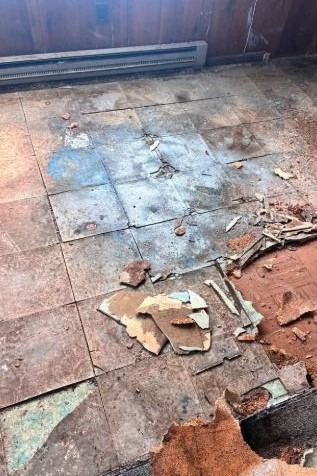 Usunięte płytki podłogowe z fragmentami gruzu i resztkami kleju na podłodze. Widoczny grzejnik przy ścianie z boazerią. Ujęcie z góry, ukazujące stan po demontażu.