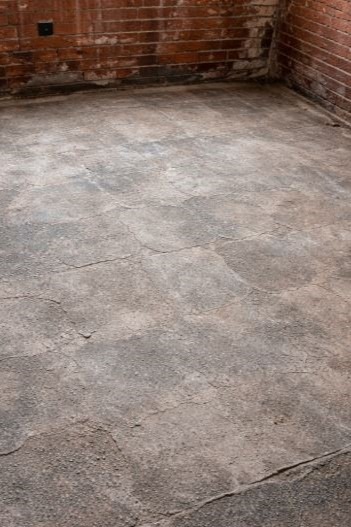 Szara, popękana posadzka w pomieszczeniu z fragmentem ceglanej ściany. Widoczne ślady po usuniętych elementach na ścianie. Surowe wykończenie wnętrza.