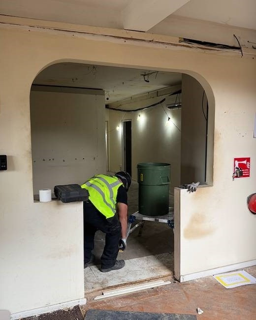 Pracownik w kamizelce przy wyburzonym łuku ściany, widoczne narzędzia i pojemnik na odpady. Ślady po usuniętej ścianie, surowy stan wnętrza.