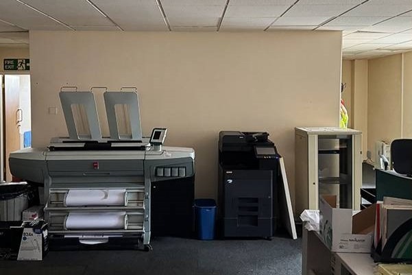 Wnętrze biura z dużym ploterem, drukarką i szafką. Widoczny kosz na śmieci i kartony. Ściany w kolorze beżowym, sufit biały z widocznymi kablami.