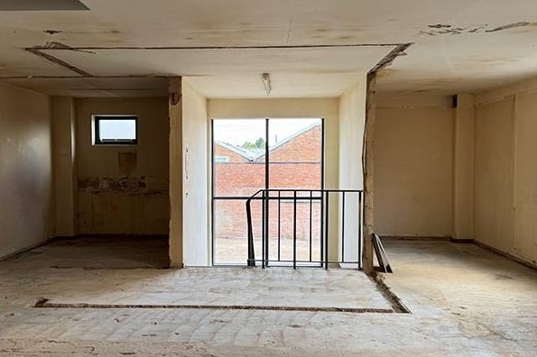 Wnętrze po wyburzeniu ścian działowych, widoczne ślady po instalacjach w podłodze i suficie, okno z widokiem na ceglany budynek. Prace rozbiórkowe w trakcie.