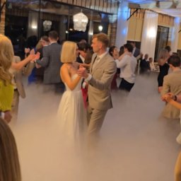 Best Event Wojciech Kulikowski - Para młoda tańczy pierwszy taniec w dymie na sali weselnej z dekoracjami świetlnymi. W tle goście i drewniana konstrukcja dachu.