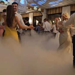 Best Event - Tańczący goście weselni w dymie, oświetleni dekoracyjnymi światłami pod sufitem namiotu. Widoczne drewniane elementy konstrukcyjne i białe zasłony.