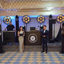 Best Event - DJ Wojtecki prezentuje nagłośnienie i oświetlenie na tle dekoracji sali. Widoczni prezenterzy w eleganckich strojach i sprzęt DJ-ski.