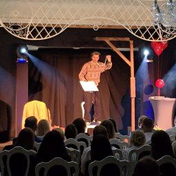 Best Event - Iluzjonista Pan Li na scenie z czarnym tłem, oświetleniem i dekoracjami balonowymi, prezentuje program przed publicznością siedzącą na białych krzesłach.