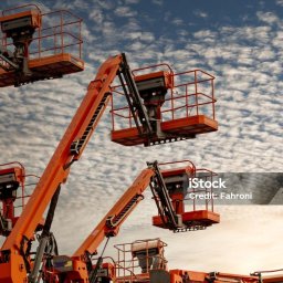 BIG Coaching Barbara Gałuszka - Pomarańczowe podnośniki koszowe na tle nieba z chmurami, prezentujące sprzęt używany podczas szkoleń operatorów maszyn budowlanych.