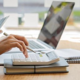 BIG Coaching Barbara Gałuszka - Kobieta używa kalkulatora, w tle laptop i notatniki. Scena sugeruje naukę finansów lub księgowości, być może w trakcie kursu zawodowego.