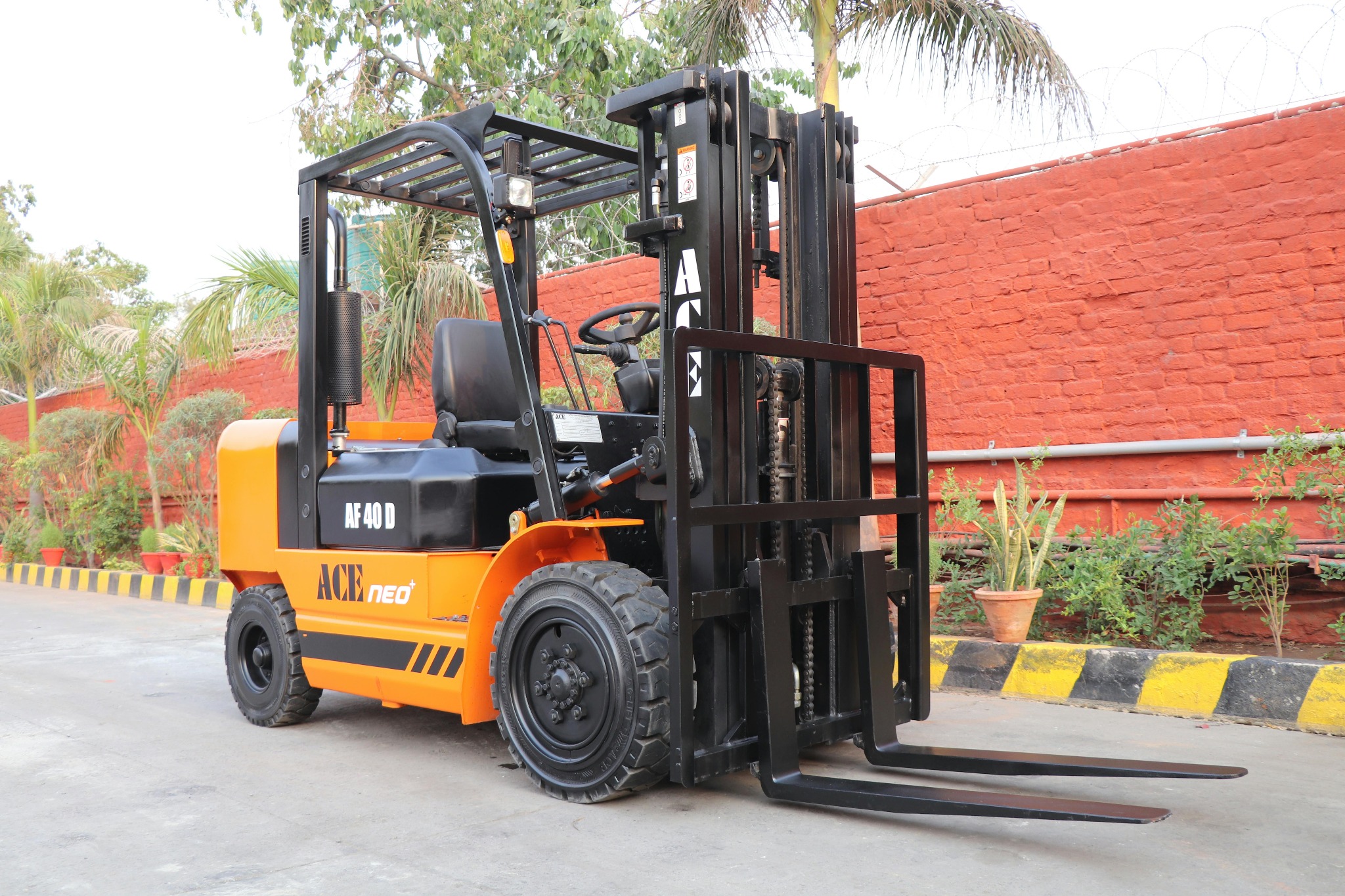 Pomarańczowo-czarny wózek widłowy ACE NEO AF 40 D na tle ceglanego muru z roślinnością. Sprzęt budowlany do szkoleń operatorów.