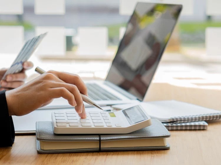 Kobieta używa kalkulatora, w tle laptop i notatniki. Scena sugeruje naukę finansów lub księgowości, być może w trakcie kursu zawodowego.