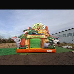 WIKTORIA WOJDA WIK-KOM - Kolorowy dmuchany zamek w stylu dżungli z motywami zwierząt, ustawiony na zielonej macie na zewnątrz, z widocznym budynkiem w tle.