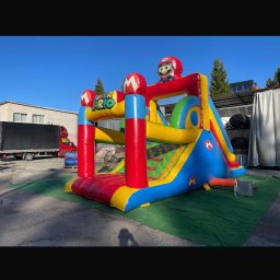 WIKTORIA WOJDA WIK-KOM - Kolorowy dmuchany zamek z motywem Mario, ustawiony na zielonej macie. W tle widoczny budynek i zaparkowany samochód dostawczy. Słoneczny dzień.