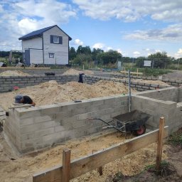 Piotr Zasadny Budownictwo - Wznoszenie fundamentów z bloczków betonowych na piaszczystym podłożu. Widoczny fragment domu w tle i pracownik przy pracy. Taczka z zaprawą obok.