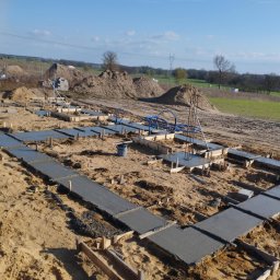 Piotr Zasadny Budownictwo - Zalane betonem fundamenty pod budowę domu, widoczne szalunki i wystające pręty zbrojeniowe. W tle hałdy piasku i elementy infrastruktury.