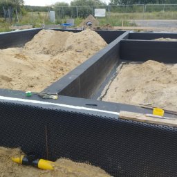 Piotr Zasadny Budownictwo - Fundamenty domu w trakcie budowy: ściany fundamentowe zabezpieczone folią kubełkową, wypełnione piaskiem. Widoczne narzędzia i elementy instalacji.