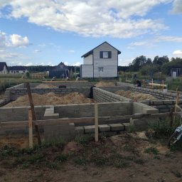 Piotr Zasadny Budownictwo - Fundamenty z bloczków betonowych w trakcie budowy, widoczne zbrojenia i piasek. W tle dom w budowie i inne zabudowania. Na pierwszym planie rower dziecięcy.