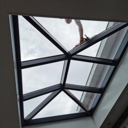 Piotr Zasadny Budownictwo - Montaż okna dachowego z czarną ramą, widok od dołu. Widoczne narzędzia i ręka montera. Przez okno widać pochmurne niebo. Ujęcie z dołu, podkreślające geometrię konstrukcji.
