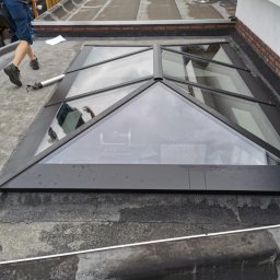 Piotr Zasadny Budownictwo - Montaż okna dachowego w kształcie piramidy na płaskim dachu pokrytym papą. Widoczny pracownik i narzędzia. Nietypowy kształt okna.