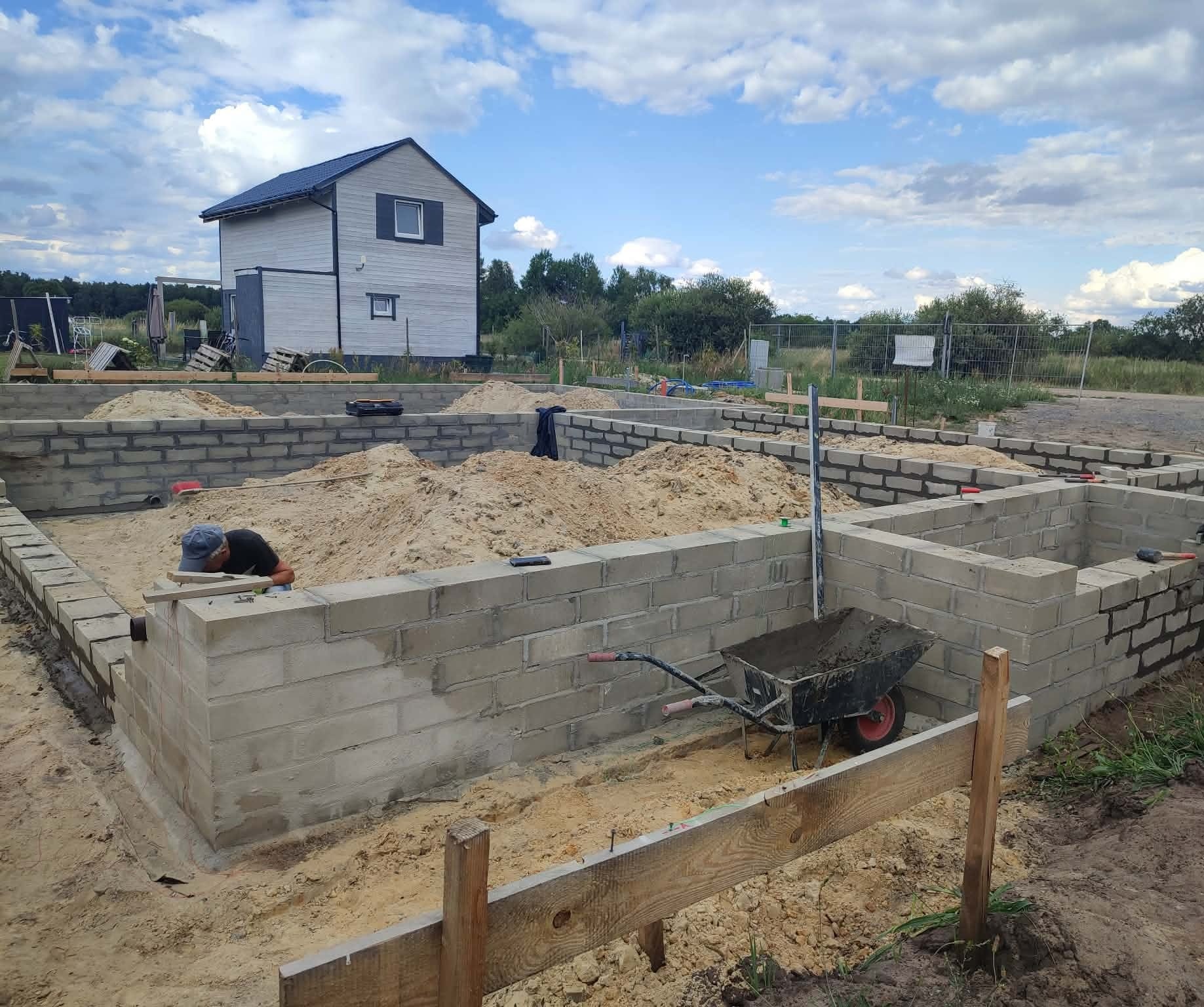 Wznoszenie fundamentów z bloczków betonowych na piaszczystym podłożu. Widoczny fragment domu w tle i pracownik przy pracy. Taczka z zaprawą obok.