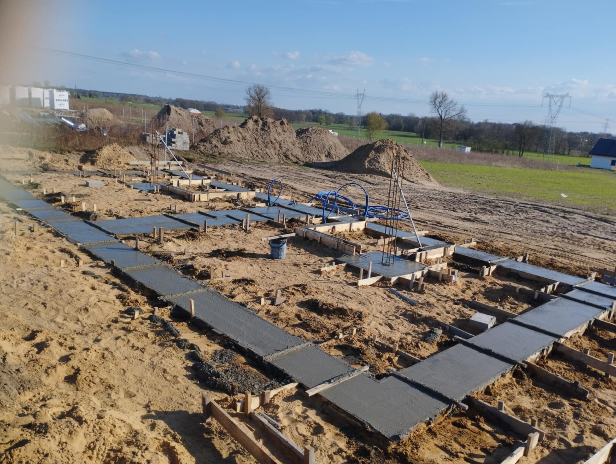Zalane betonem fundamenty pod budowę domu, widoczne szalunki i wystające pręty zbrojeniowe. W tle hałdy piasku i elementy infrastruktury.