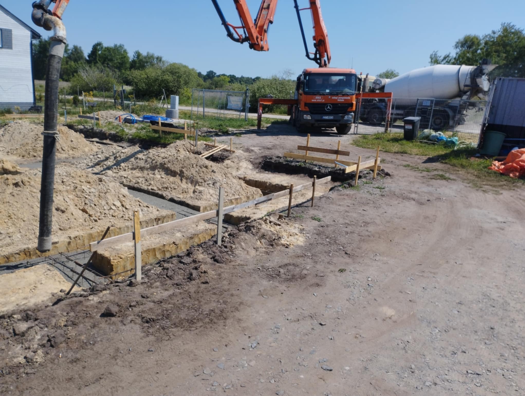 Wylewanie betonu do wykopów pod fundamenty domu jednorodzinnego. Na drugim planie betoniarka i pompa do betonu.