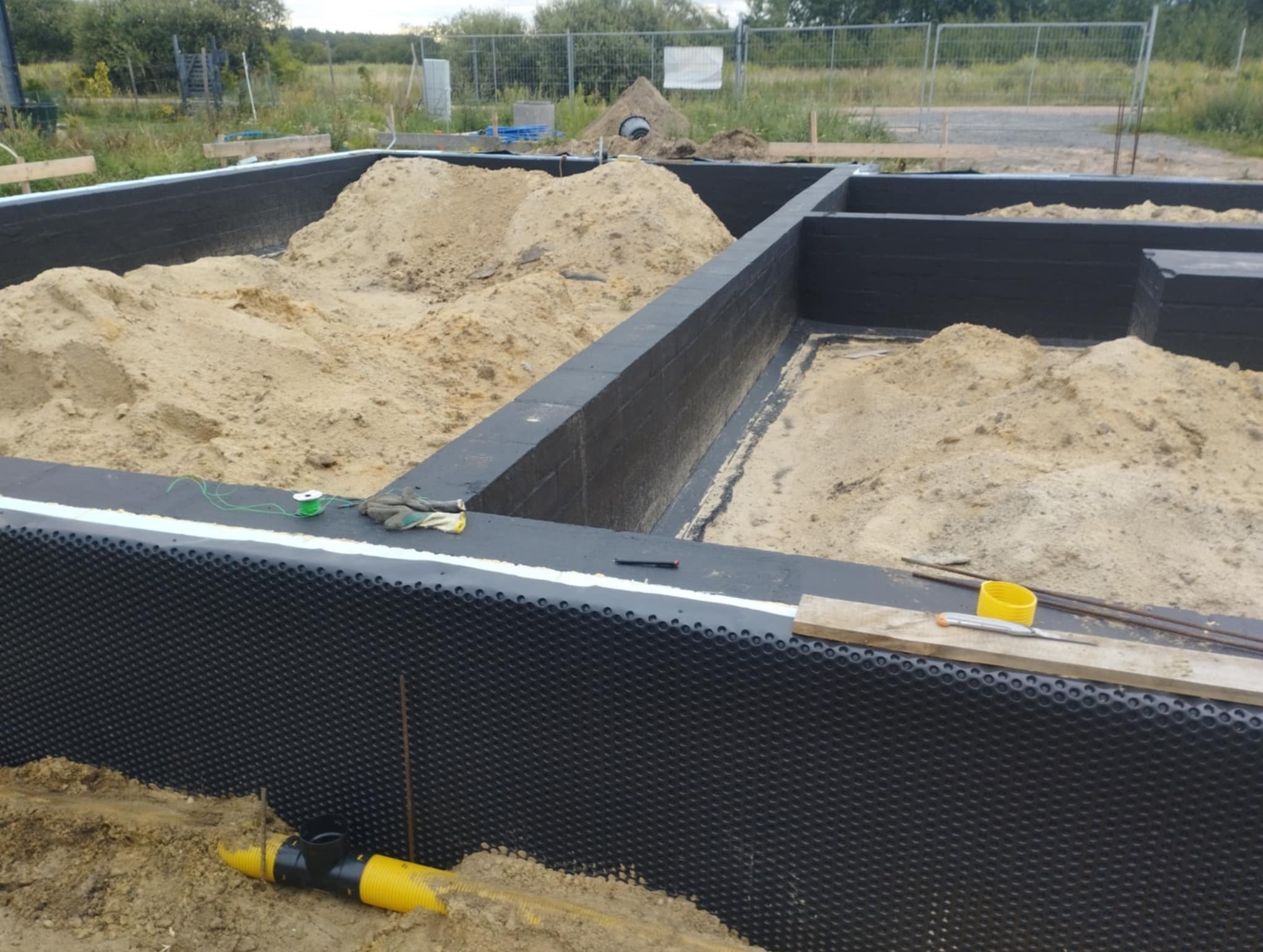 Fundamenty domu w trakcie budowy: ściany fundamentowe zabezpieczone folią kubełkową, wypełnione piaskiem. Widoczne narzędzia i elementy instalacji.