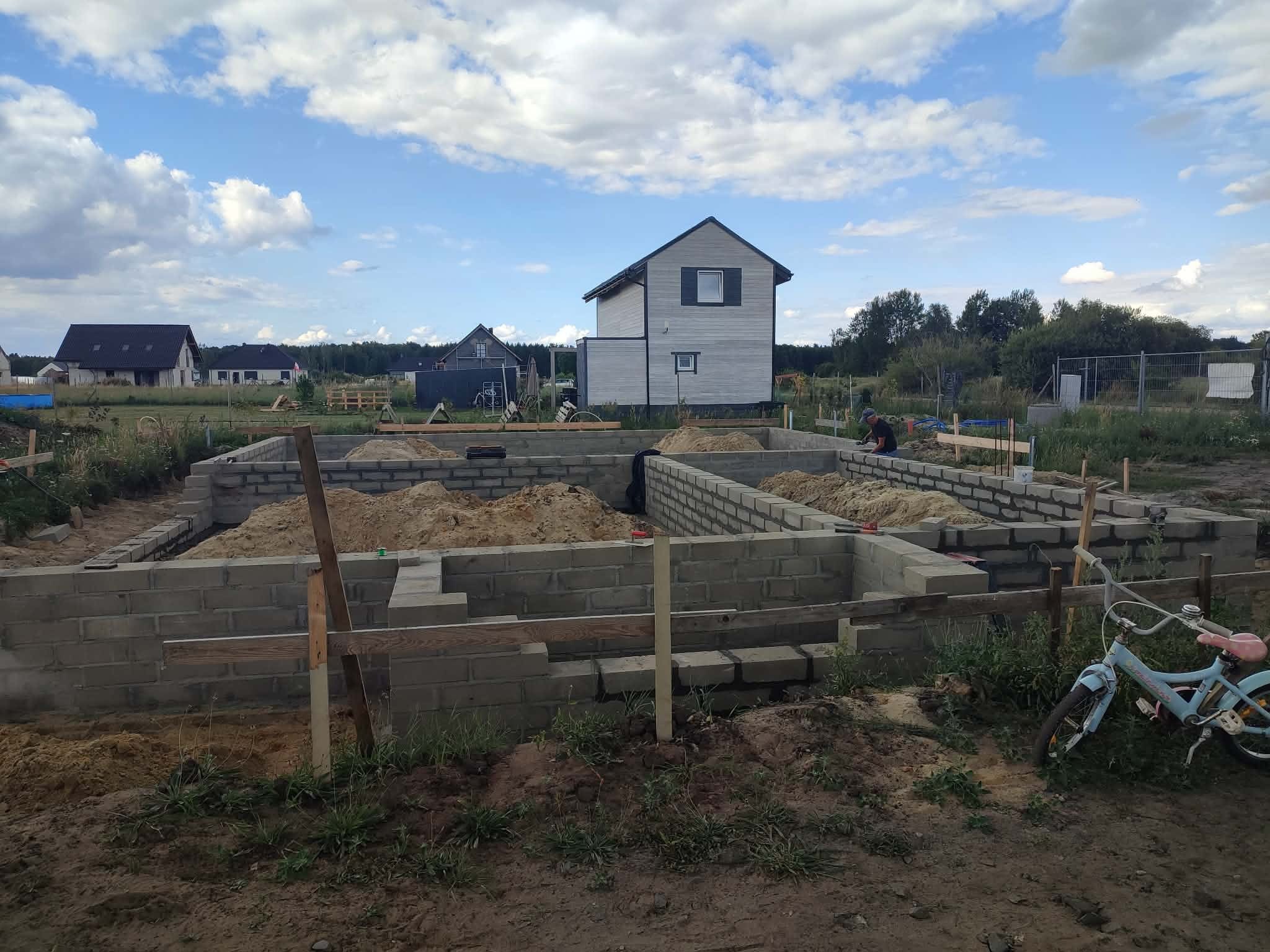 Fundamenty z bloczków betonowych w trakcie budowy, widoczne zbrojenia i piasek. W tle dom w budowie i inne zabudowania. Na pierwszym planie rower dziecięcy.
