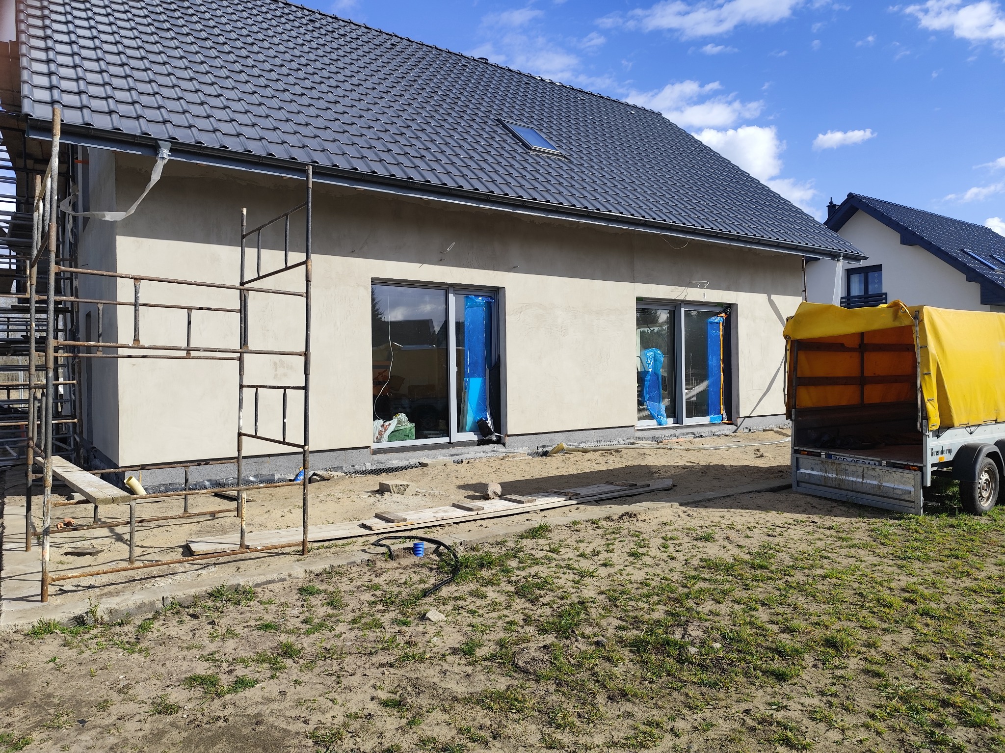 Dom w trakcie budowy: otynkowana elewacja, okna zabezpieczone folią, rusztowania, przyczepka. Widoczne fragmenty dachu z dachówką i okno dachowe.
