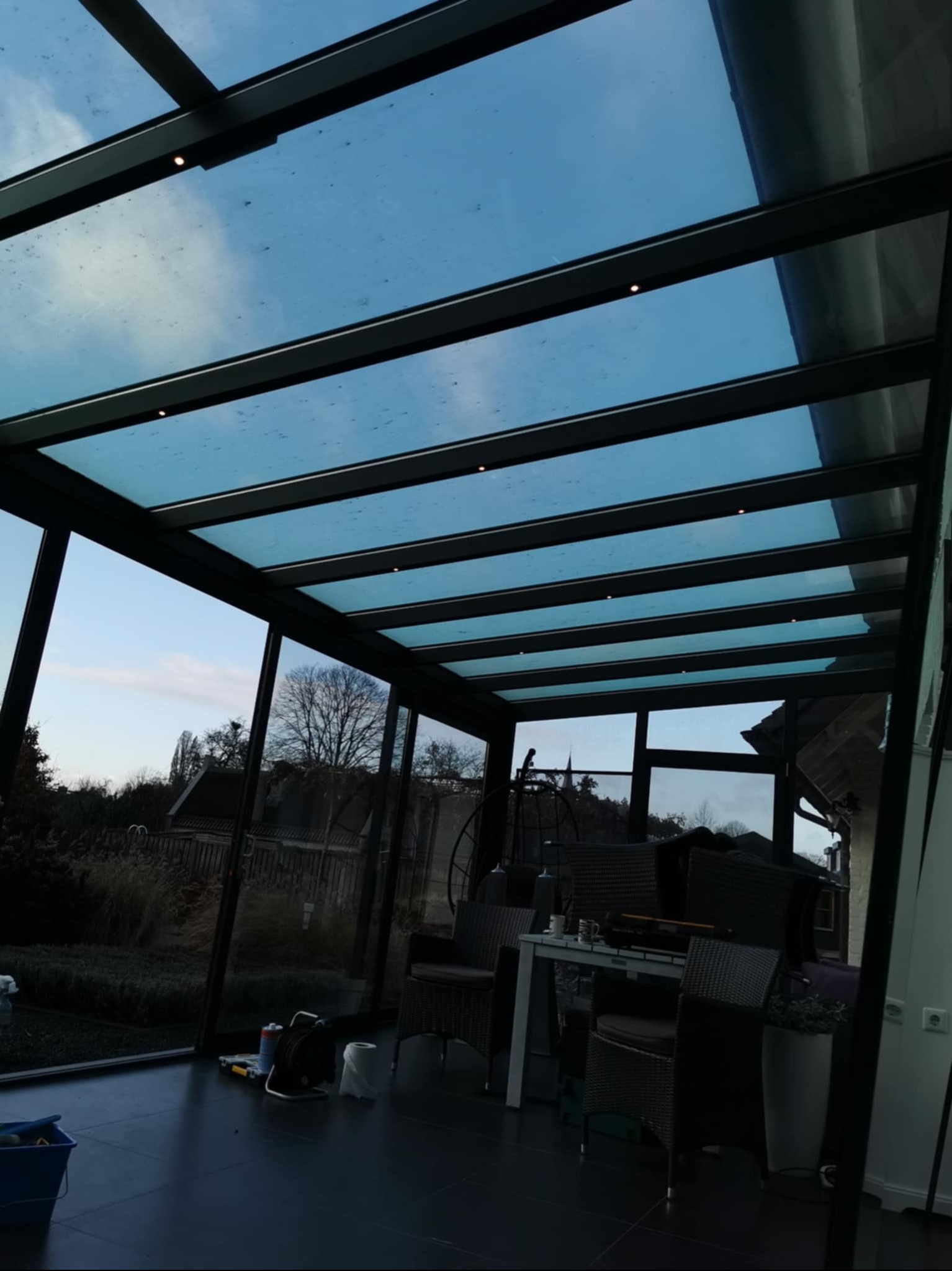 Zadaszony taras z czarną konstrukcją i przeszklonym dachem, widok na ogród. Wnętrze z meblami ogrodowymi i akcesoriami. Nowoczesny design z oświetleniem punktowym.