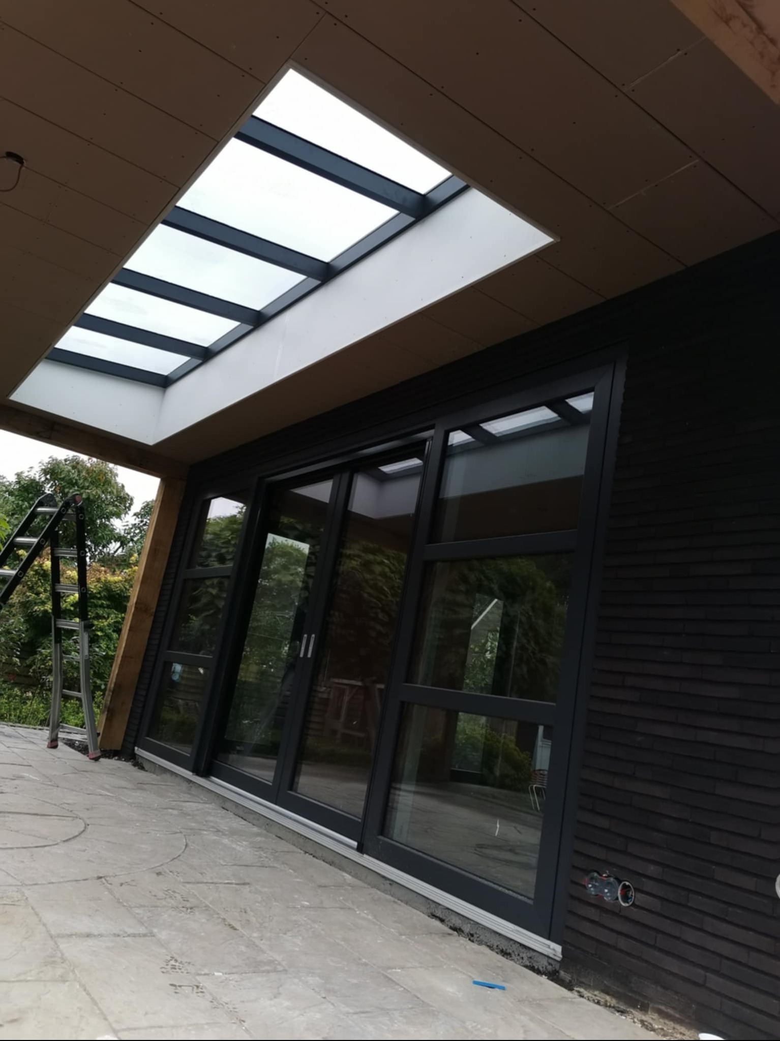 Nowoczesny taras z przesuwnymi drzwiami i świetlikiem dachowym. Ciemna elewacja budynku kontrastuje z jasną podłogą tarasu i zielenią w tle. Widoczna drabina.