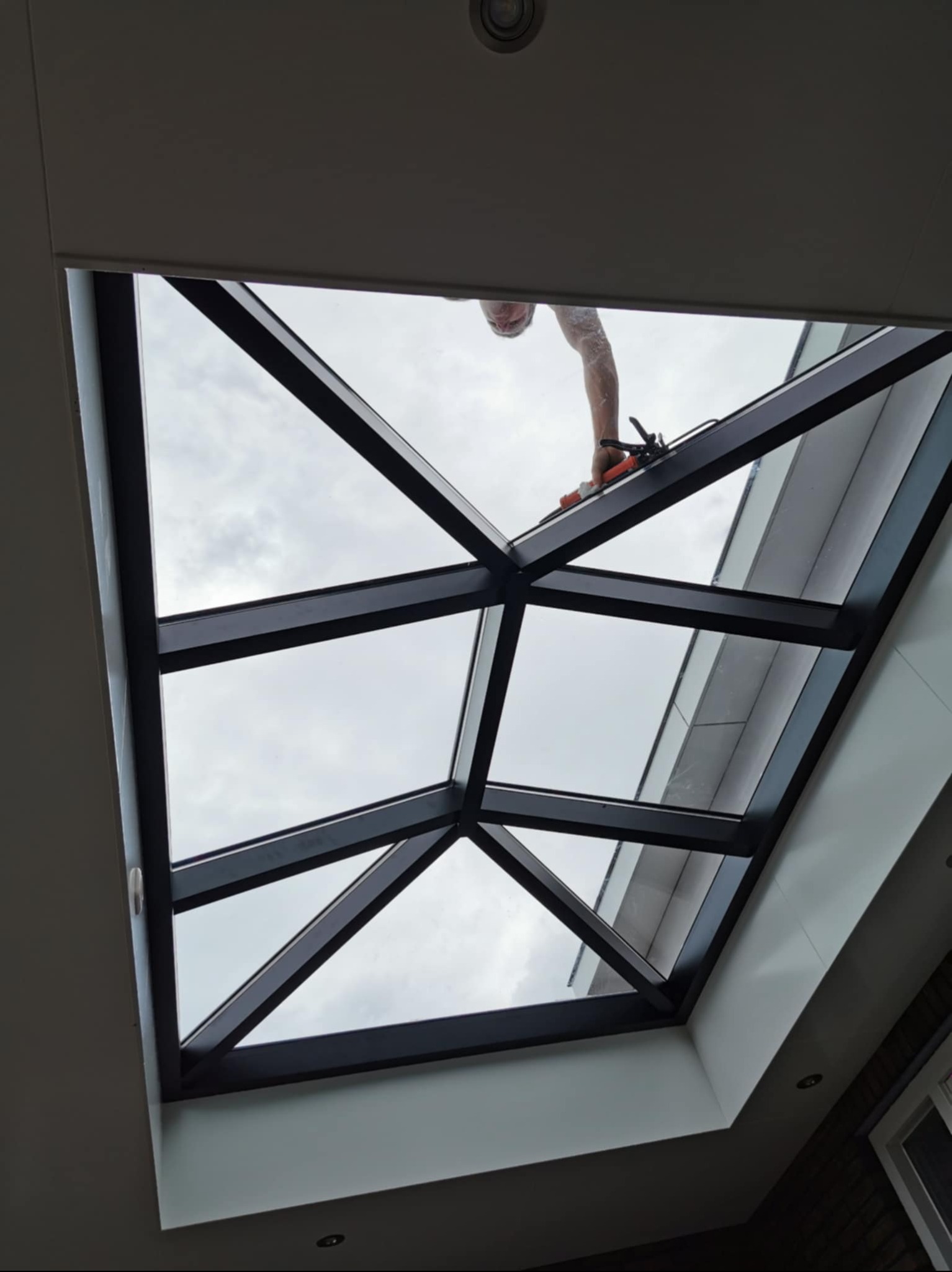 Montaż okna dachowego z czarną ramą, widok od dołu. Widoczne narzędzia i ręka montera. Przez okno widać pochmurne niebo. Ujęcie z dołu, podkreślające geometrię konstrukcji.