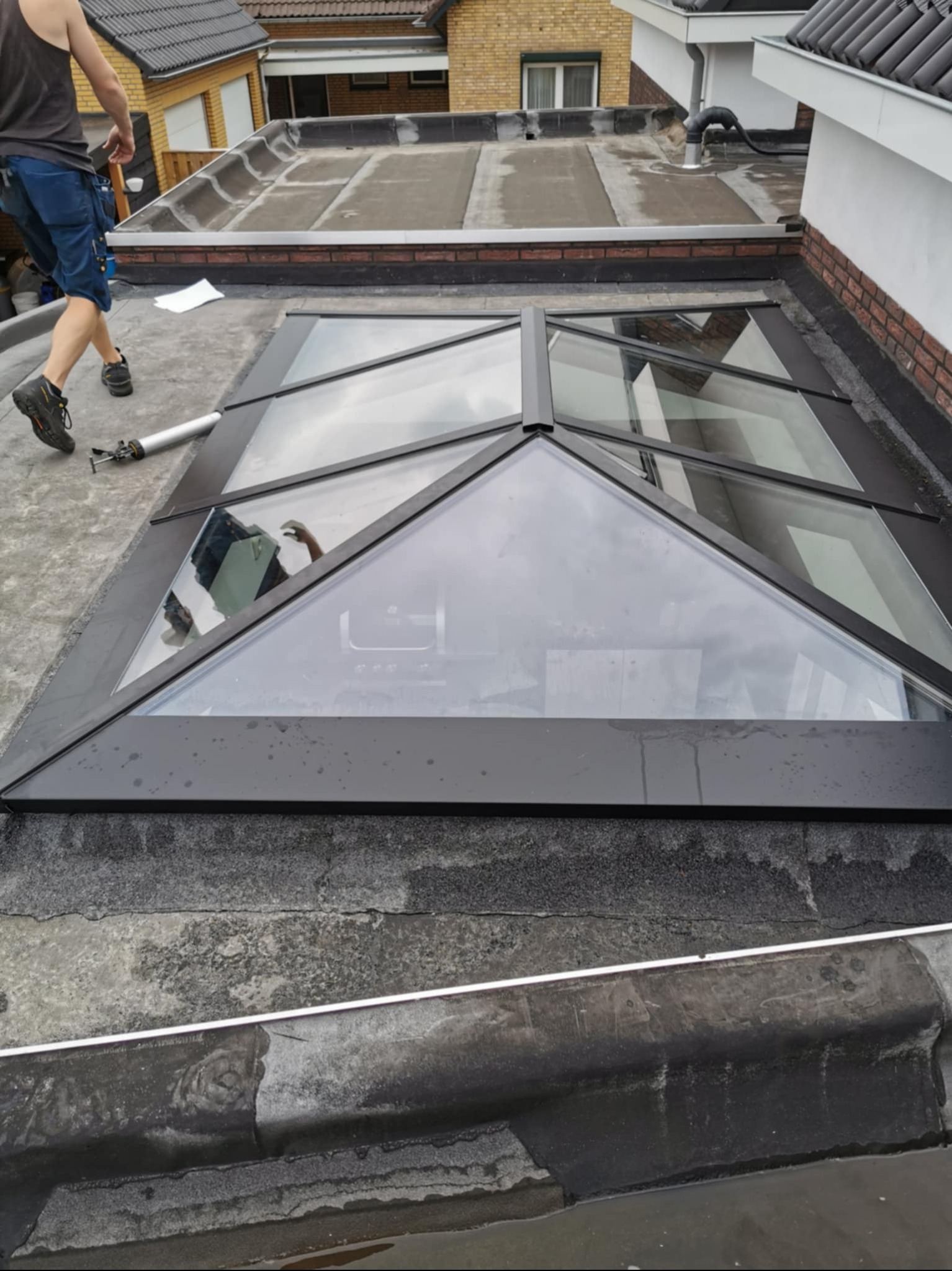 Montaż okna dachowego w kształcie piramidy na płaskim dachu pokrytym papą. Widoczny pracownik i narzędzia. Nietypowy kształt okna.