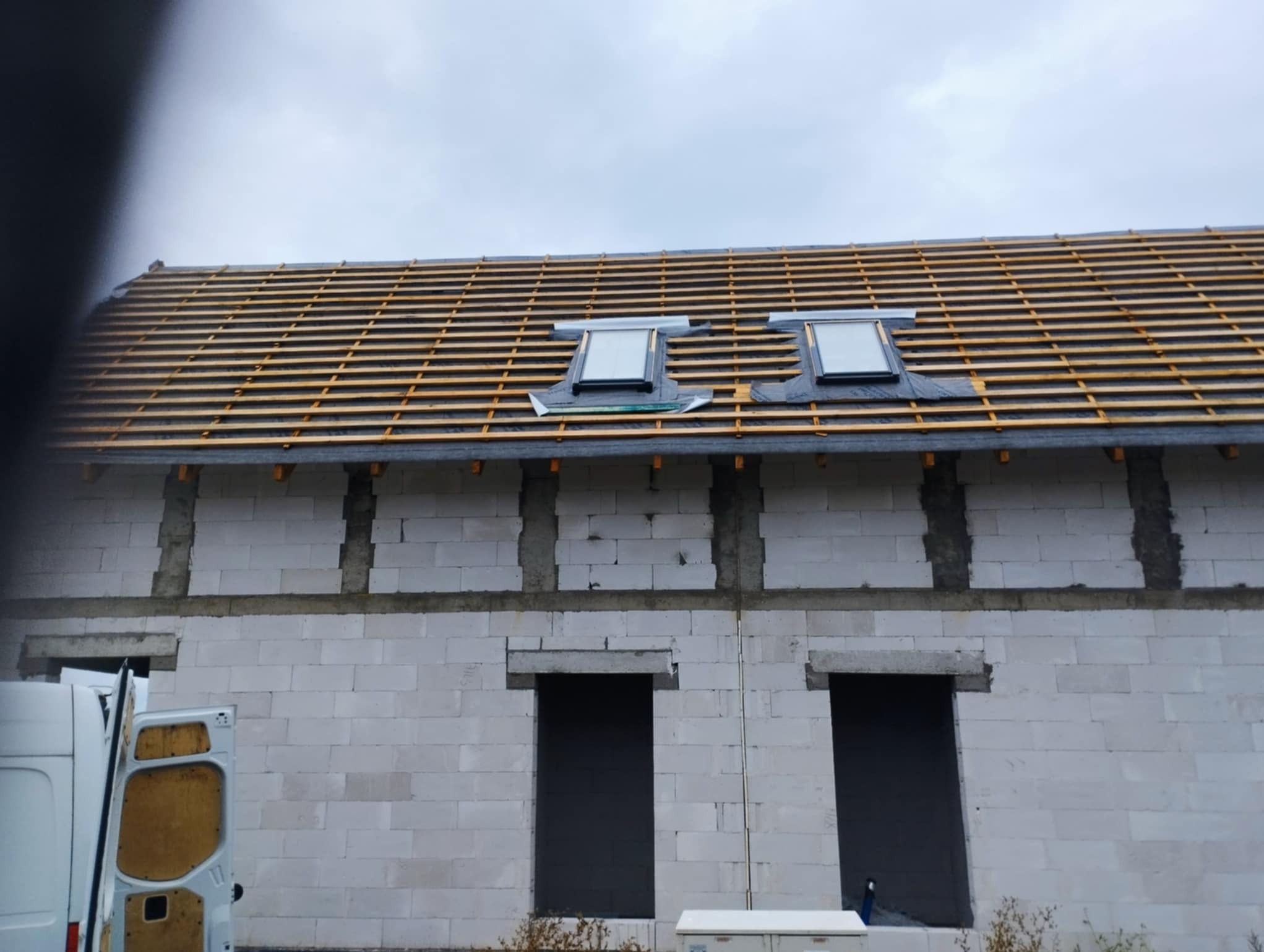 Budowa domu z pustaków: ściany, dach z łatami i dwa okna dachowe w trakcie montażu. Częściowo widoczny biały bus.