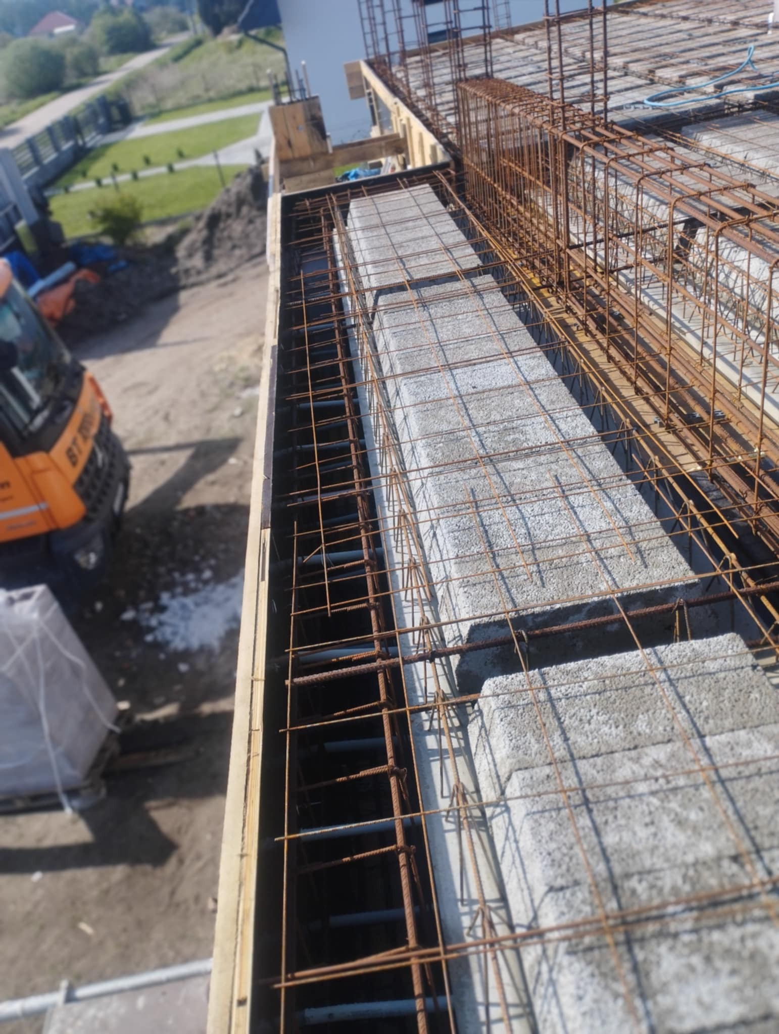 Zbrojenie stropu Teriva z pustaków szalunkowych, widoczne pręty i betonowe elementy. Na drugim planie teren budowy z koparką.