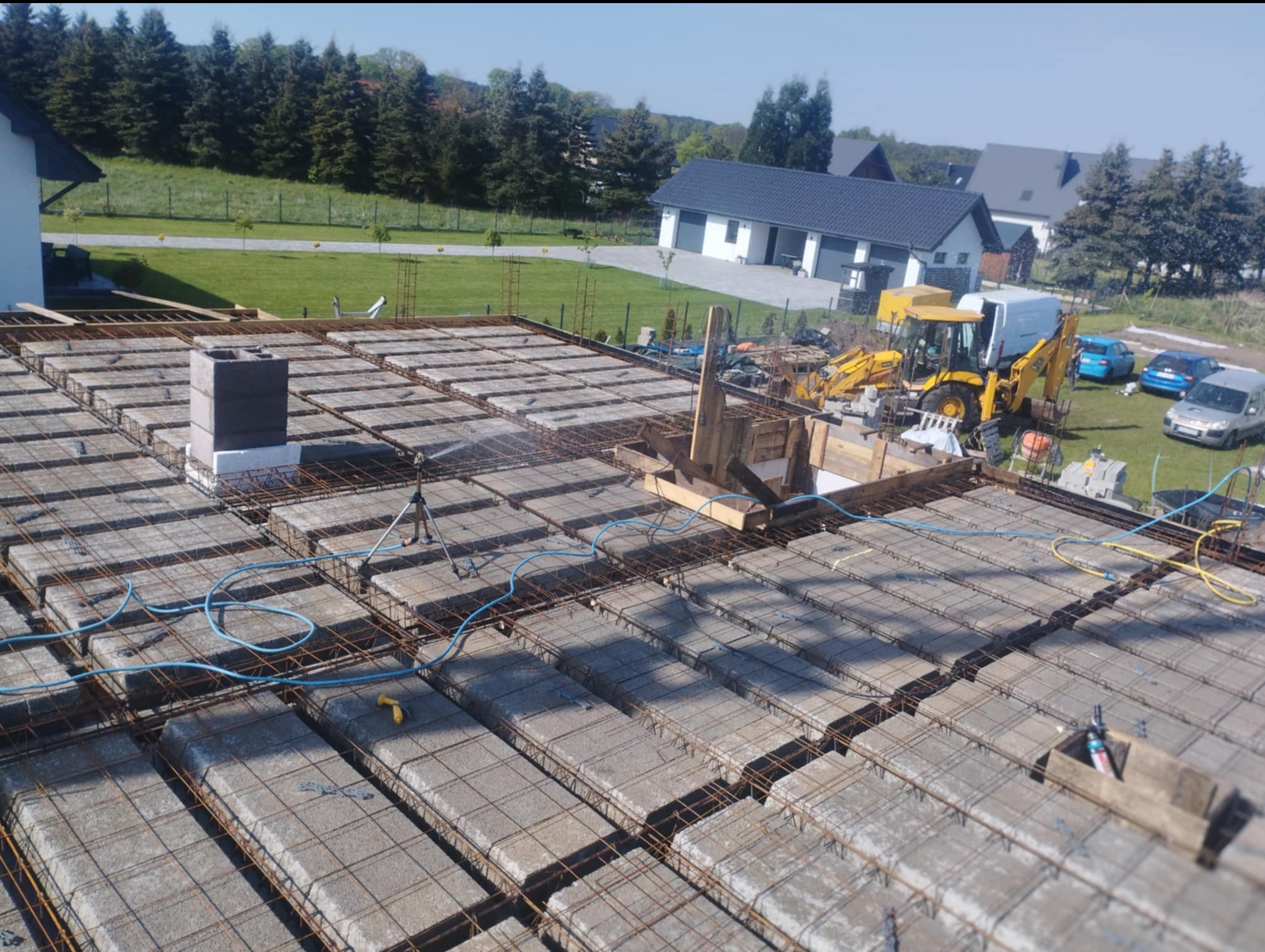 Zbrojenie stropu Teriva z pustakami betonowymi, widok z góry na budowę domu jednorodzinnego w słoneczny dzień. W tle zabudowa mieszkalna i zieleń.