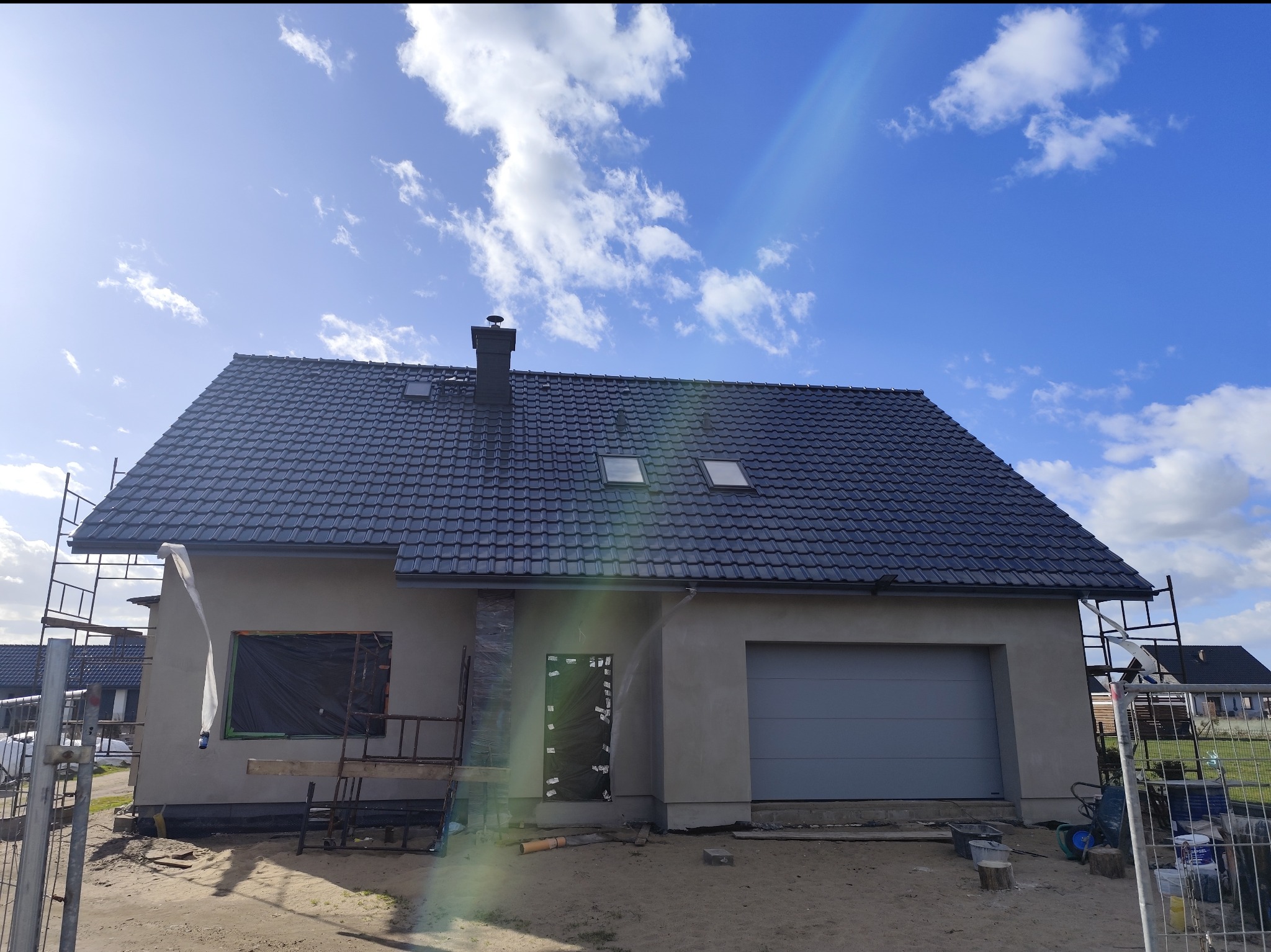 Niedokończony dom z szarą elewacją i ciemną dachówką w słoneczny dzień. Widoczne rusztowania i zabezpieczone okna. Częściowo otynkowana fasada.