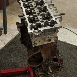 NorthTruck Norbert Kiczor - Silnik samochodowy z otwartą głowicą cylindrów, umieszczony na czerwonym stojaku warsztatowym, ukazujący mechanizm zaworowy i wnętrze silnika.