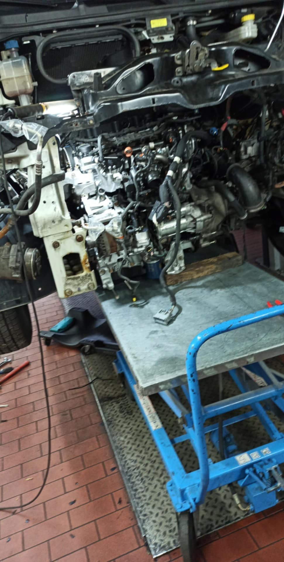 Silnik samochodowy w trakcie naprawy na podnośniku w warsztacie. Widoczne przewody, elementy mechaniczne i narzędzia. Ceglana podłoga i metalowa platforma.