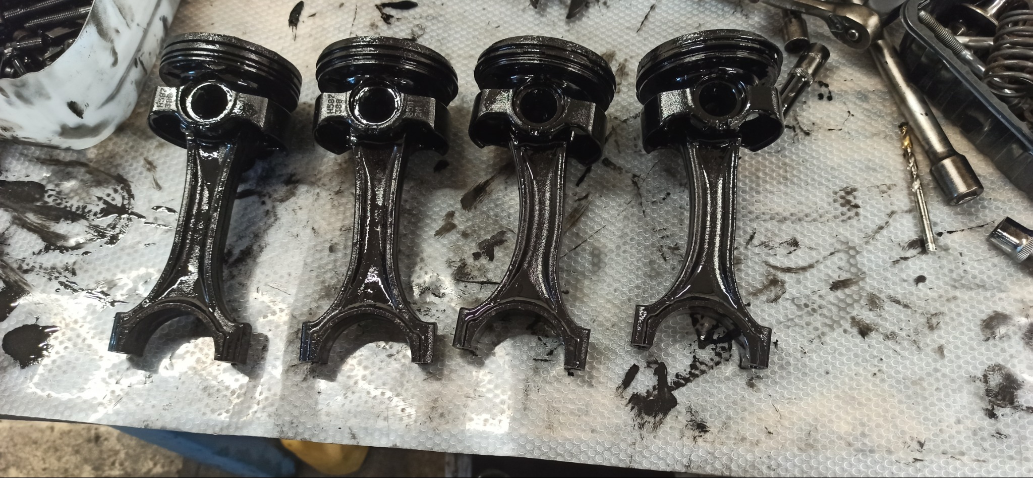 Cztery brudne tłoki silnika ułożone na warsztatowym stole, widoczne ślady oleju i narzędzia, co sugeruje etap naprawy lub diagnostyki.