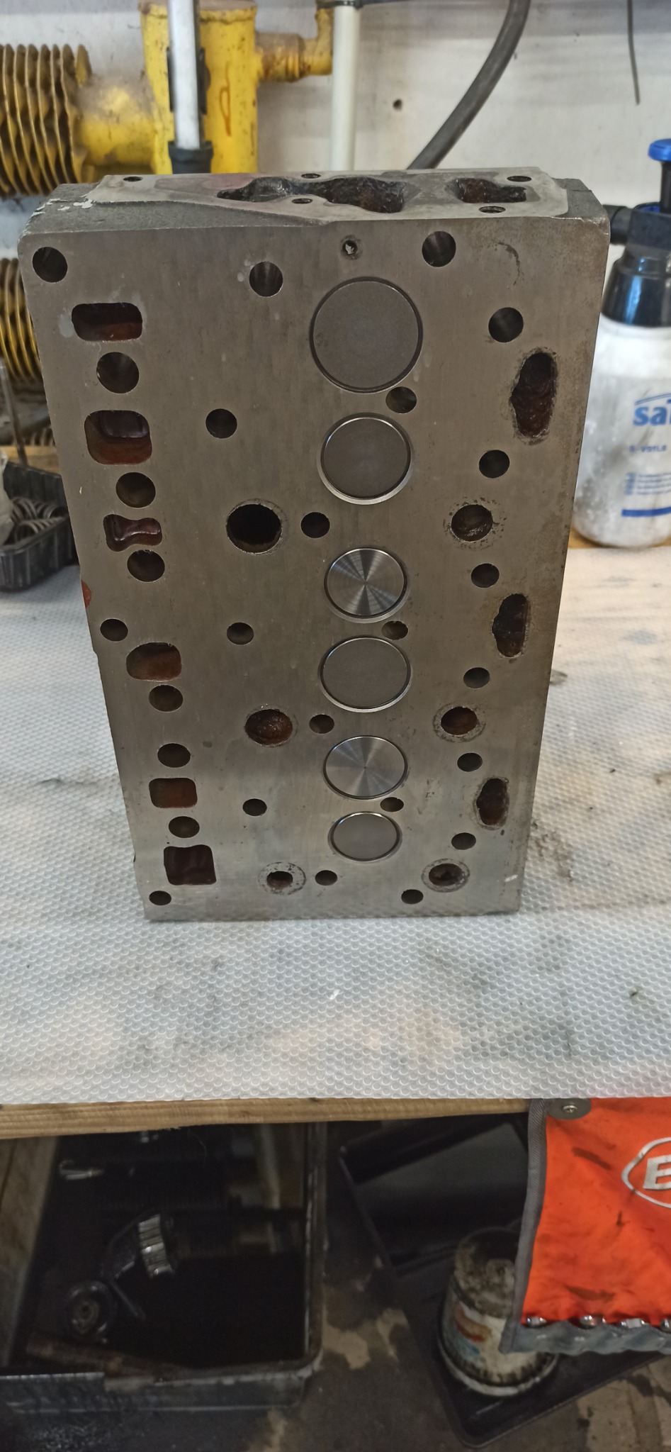 Głowica silnika spalinowego z widocznymi zaworami, osadzona na warsztatowym stole. Widoczne ślady użytkowania i korozji.