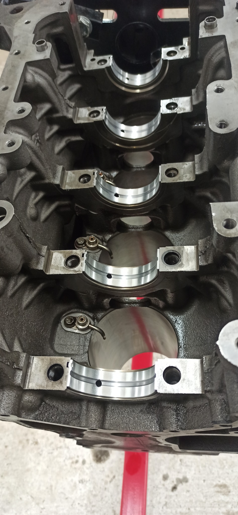 Blok silnika z zamontowanymi panewkami głównymi, widoczne ślady oleju, uchwyty panewek z zabezpieczeniami. Detal z warsztatu samochodowego.