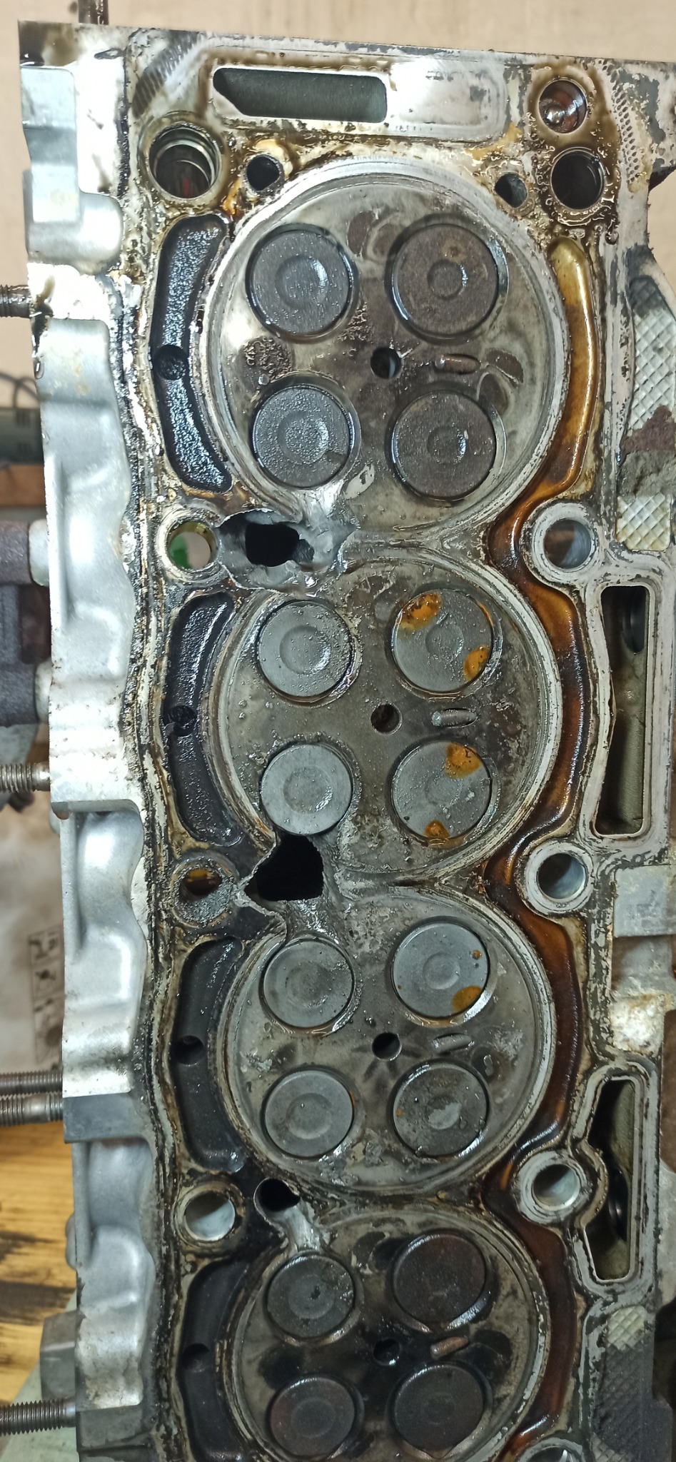 Uszkodzona głowica silnika z widocznymi zaworami, nagarem i śladami oleju. Widoczne pęknięcia i korozja. Detal z bliska, ukazujący zużycie.