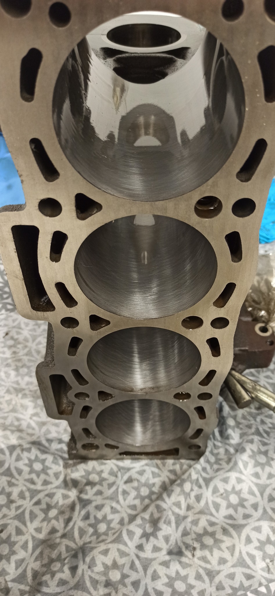 Blok silnika z trzema cylindrami po honowaniu, widoczne ślady obróbki. Otwory technologiczne w korpusie. Tło stanowi wzorzysta tkanina.