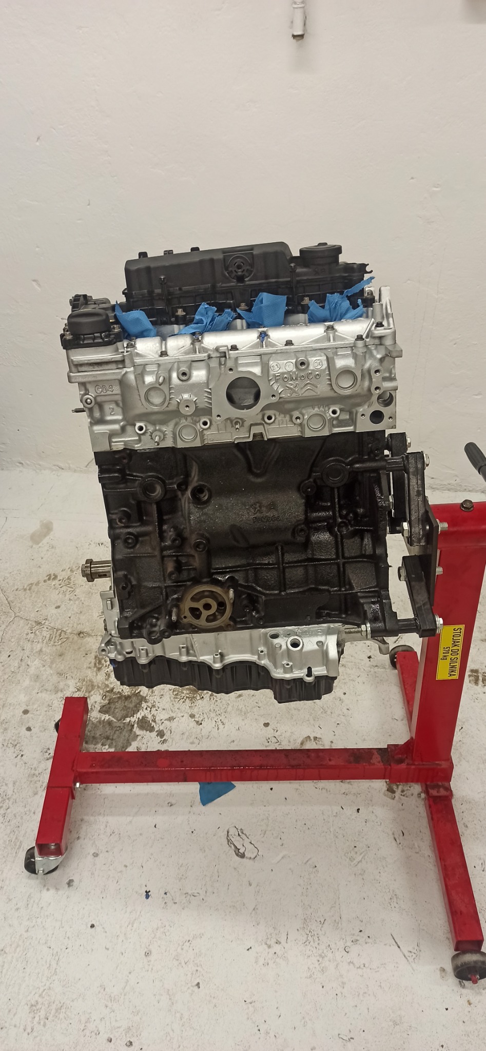 Silnik samochodowy na czerwonym stojaku warsztatowym, częściowo odnowiony, z widocznymi śladami użytkowania i świeżym lakierem na górnej części bloku.