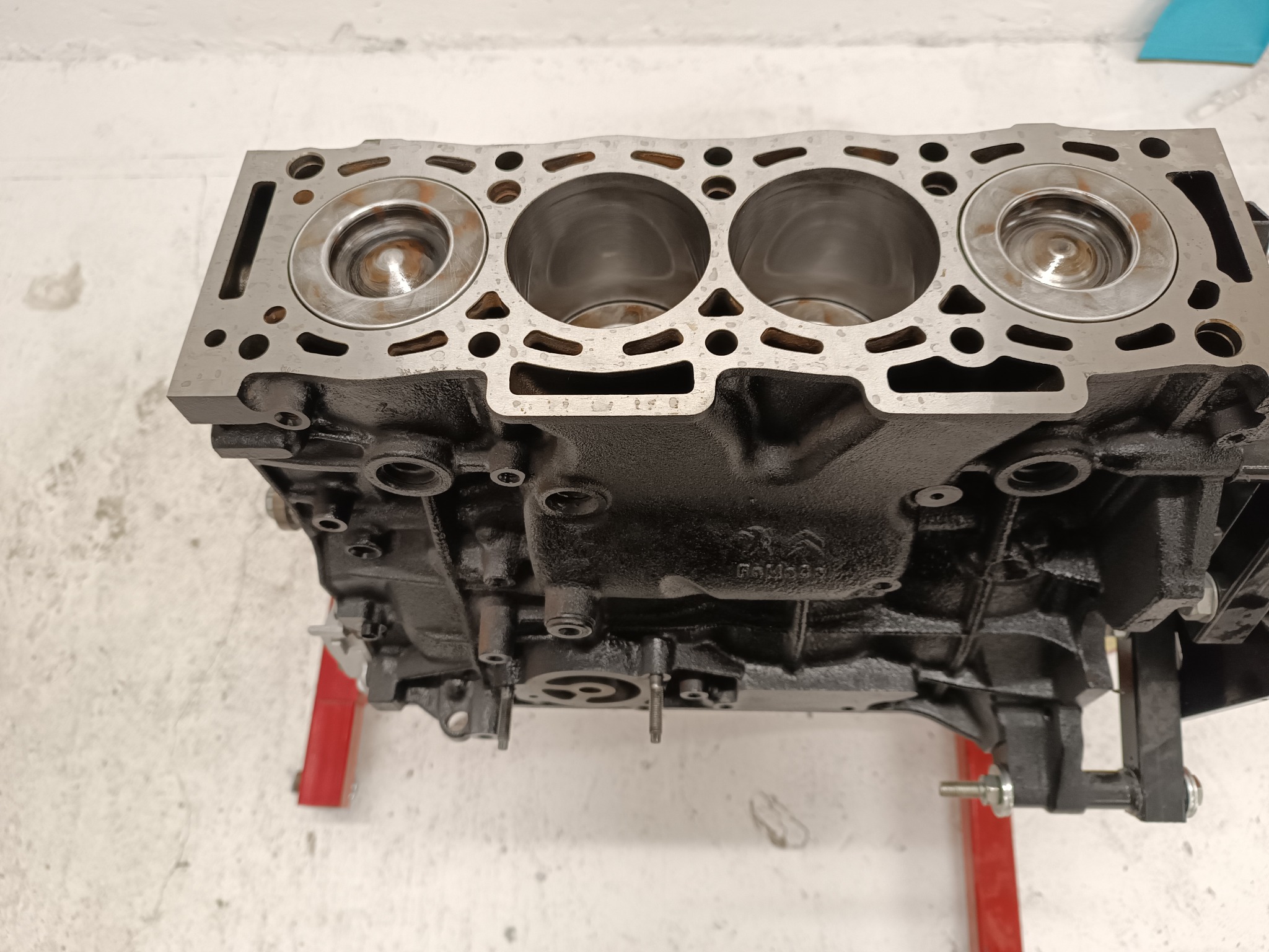 Czarny blok silnika z trzema cylindrami, widoczne tłoki, umieszczony na czerwonym stojaku warsztatowym na tle białej ściany.