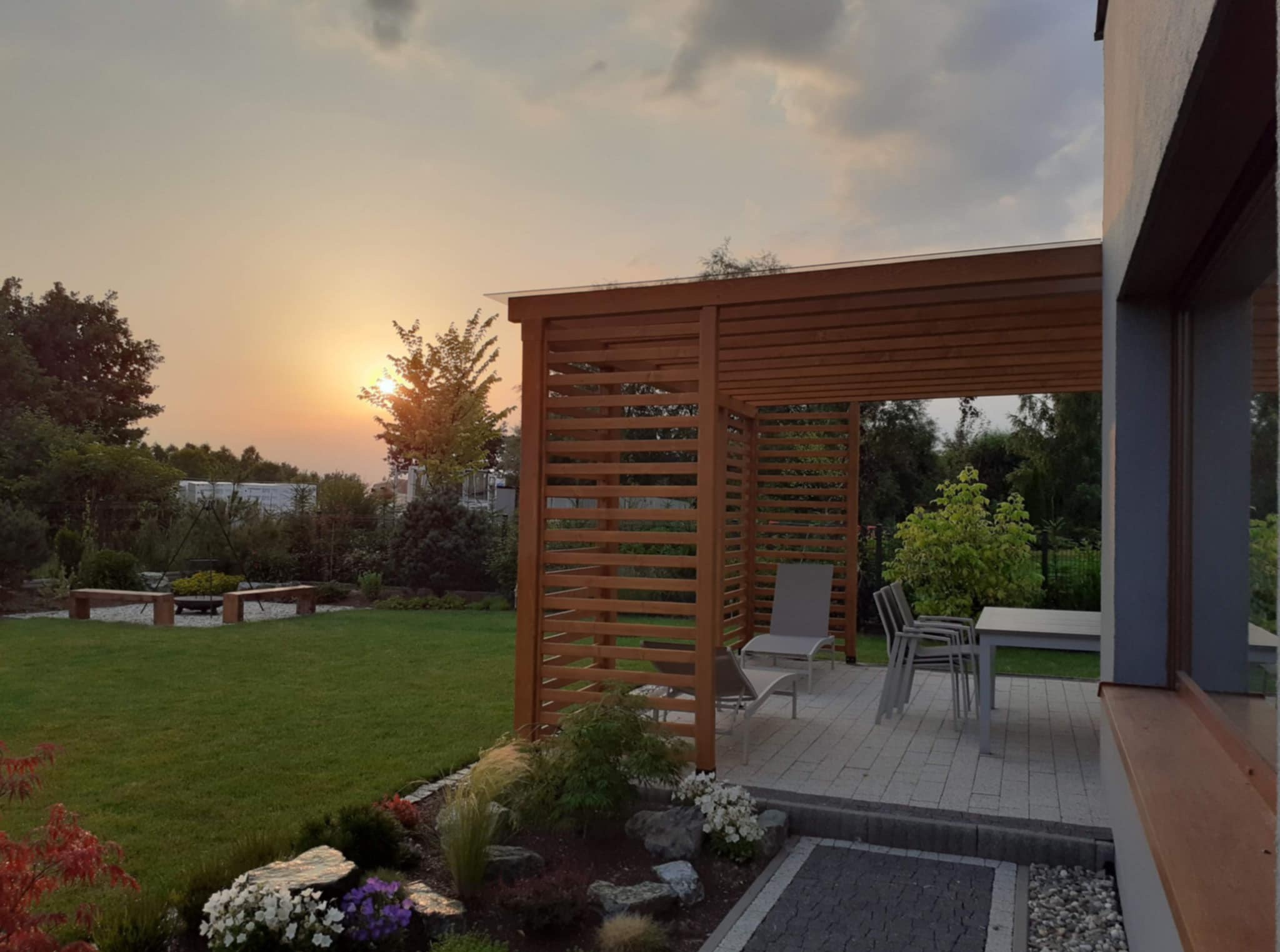 Drewniana pergola z meblami ogrodowymi na tle zachodzącego słońca. Widok na zadbany trawnik i elementy architektury ogrodowej. Ciepłe światło podkreśla naturalny charakter.