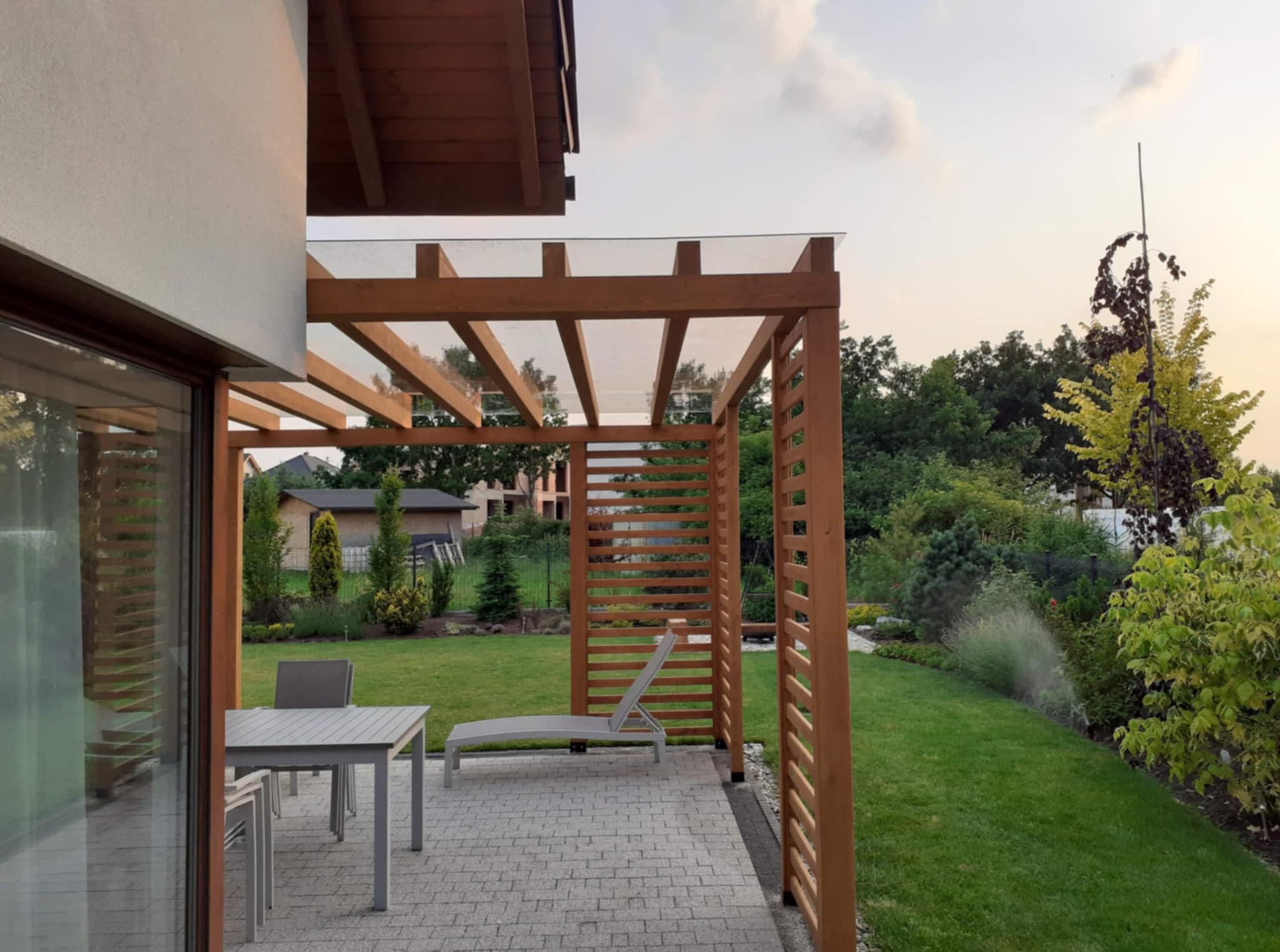 Drewniana pergola z przezroczystym zadaszeniem, szary stół z krzesłami i leżak na brukowanym tarasie przylegającym do domu. W tle zielony trawnik i drzewa.