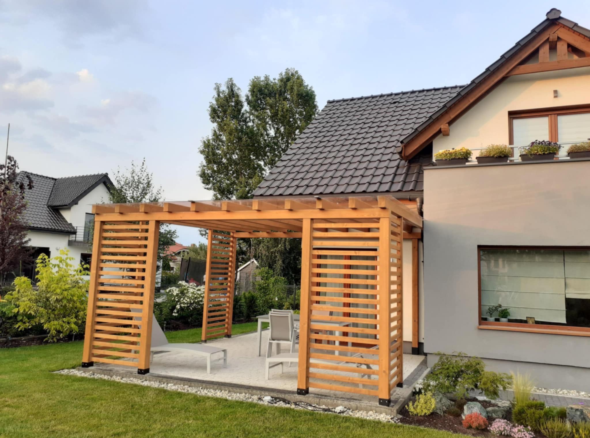 Drewniana pergola z ażurowymi ścianami, z jasnego drewna, na tle domu z szarą elewacją i zielonym trawnikiem. Widoczne meble ogrodowe.