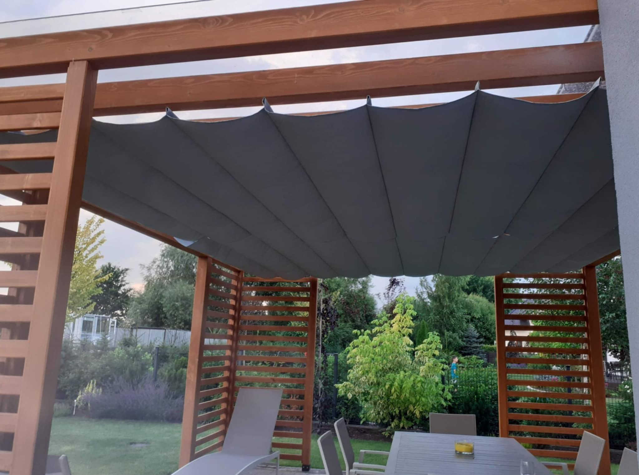 Drewniana pergola z rozsuwanym zadaszeniem materiałowym, widok z wnętrza na ogród. Meble ogrodowe pod pergolą. Konstrukcja z belek i listew w ciepłym odcieniu brązu.