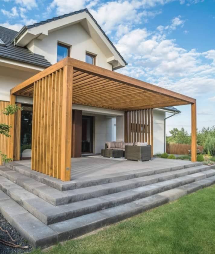 Drewniana pergola z ażurowymi ściankami na tarasie w Bulowicach. Schody z szarej kostki prowadzą do jasnego domu z ciemnym dachem. Meble ogrodowe na tarasie.